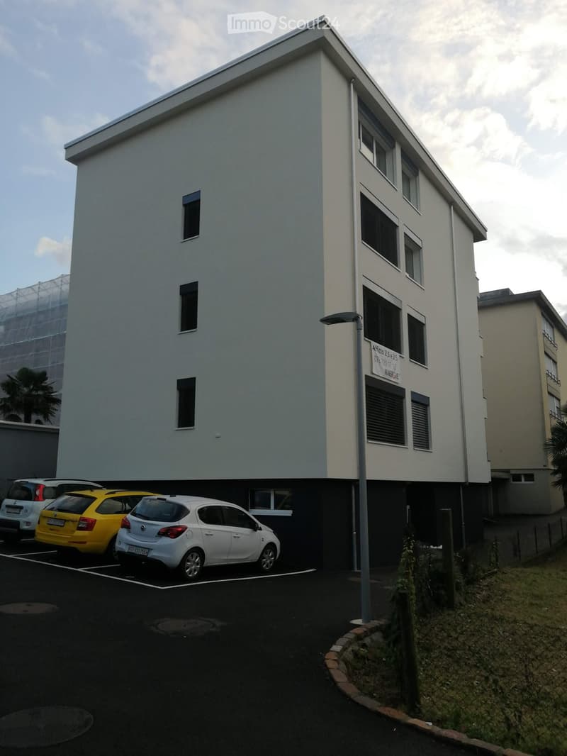 Modern multi-story building with white facade and multiple windows, located next to a parking area with several cars.