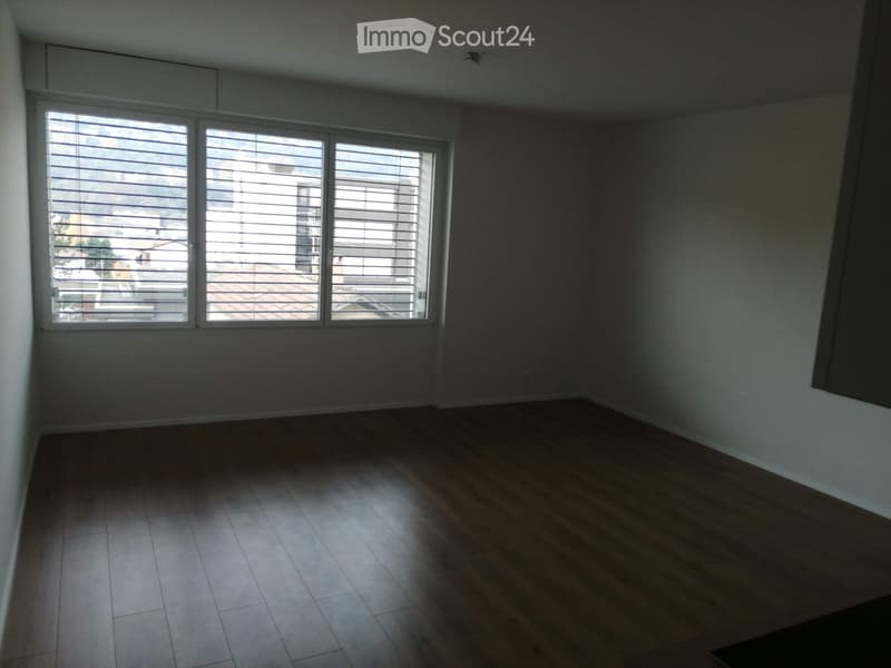 Empty room with wooden flooring and large windows with blinds, offering a view of the outside area.