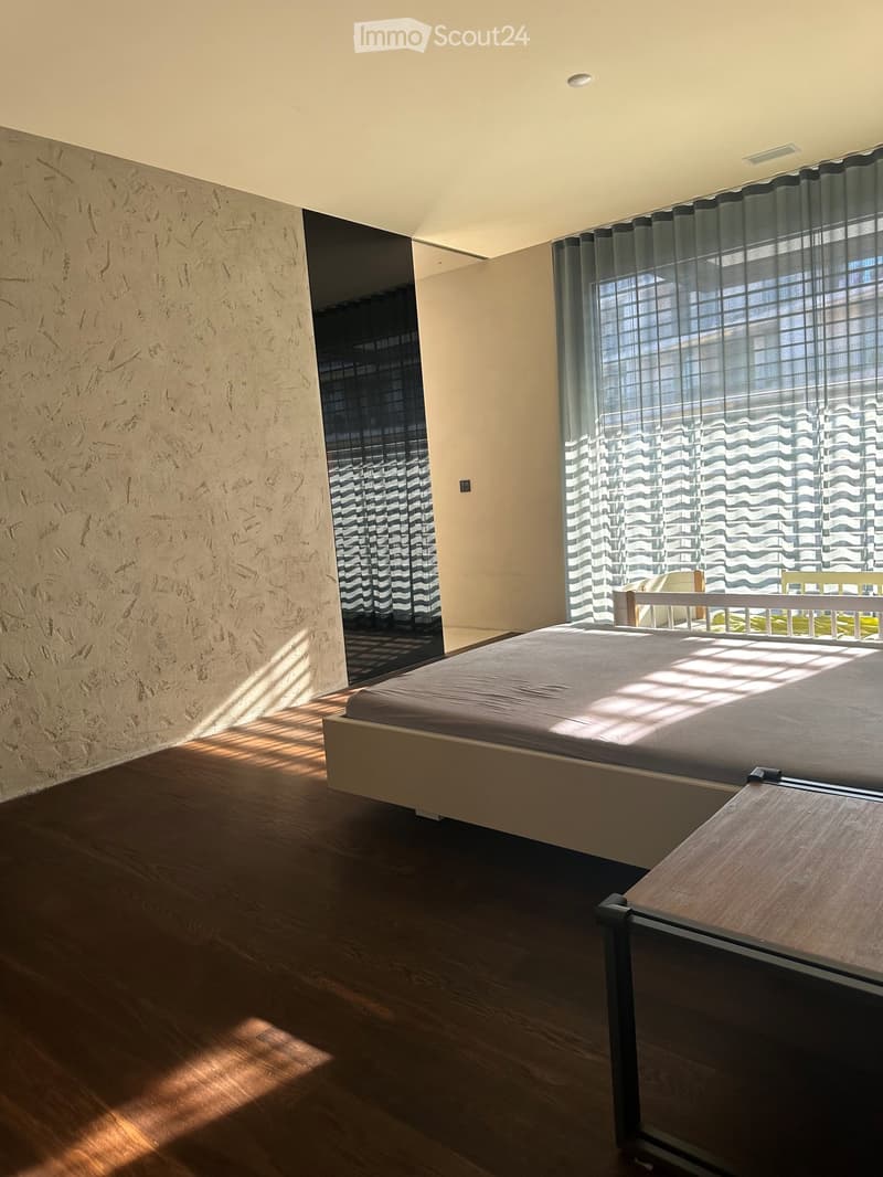 Modern bedroom with wooden flooring, large window with sheer curtains, bed with dark bedding, nightstand, and chair visible.