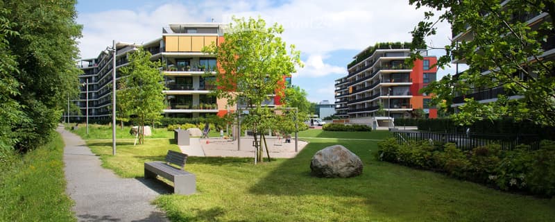A well-maintained park area with a bench, trees, and a large rock, surrounded by modern apartment buildings.