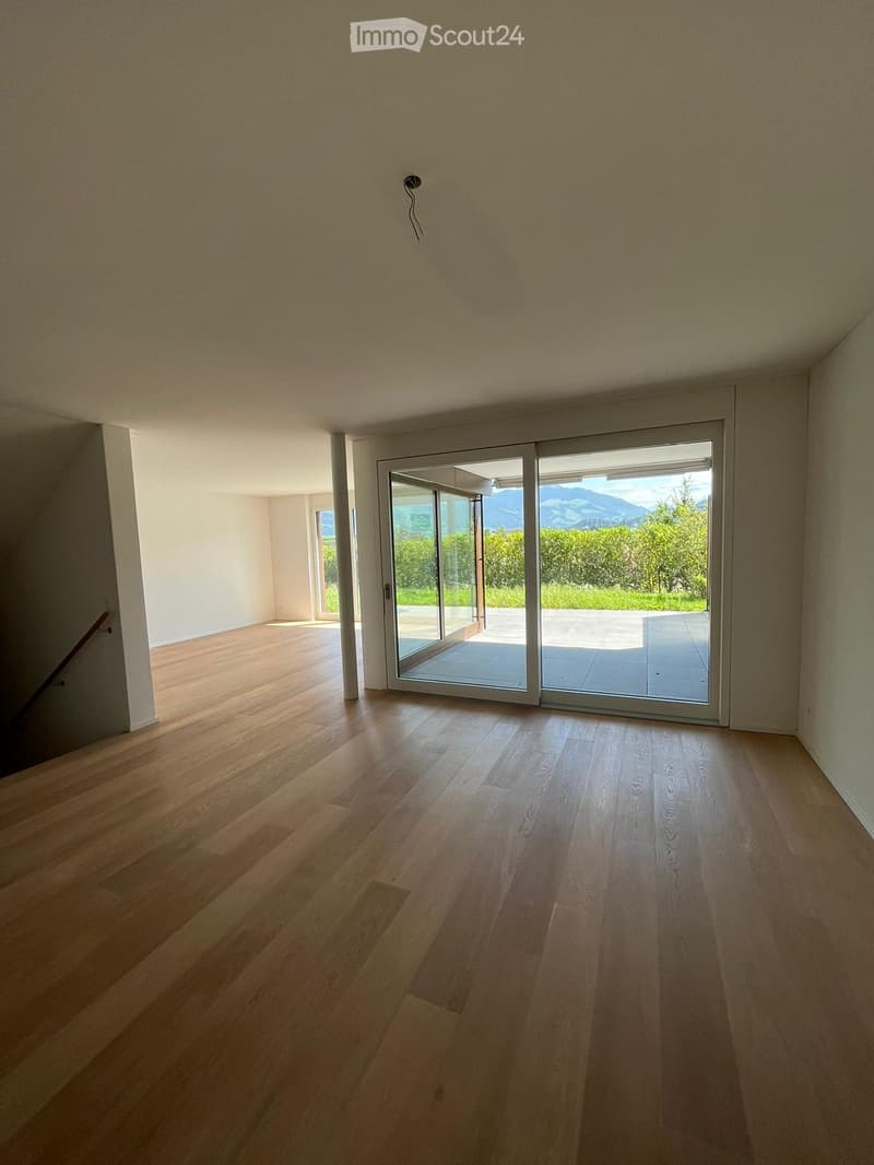 Empty room with light wooden flooring, white walls, and large sliding glass doors leading to an outdoor area with a view of greenery and mountains.