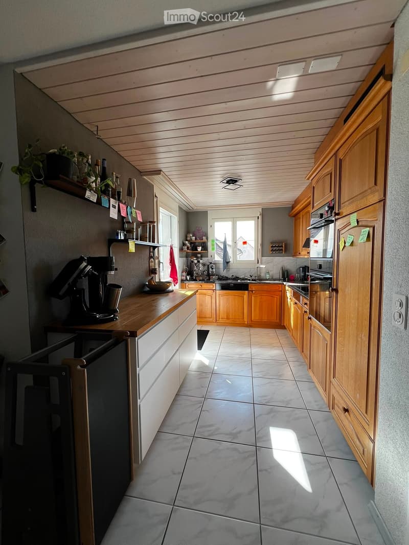 Modern kitchen with wooden cabinets, tiled floor, and various appliances including a refrigerator, oven, dishwasher, and coffee maker. Natural light streams in from windows.