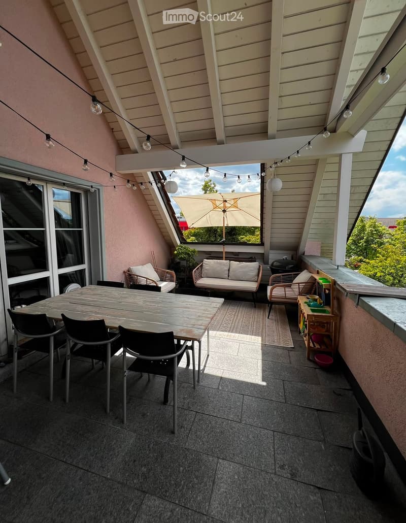 Balcony with wooden table, chairs, sofa, umbrella, and storage unit, overlooking a green area.