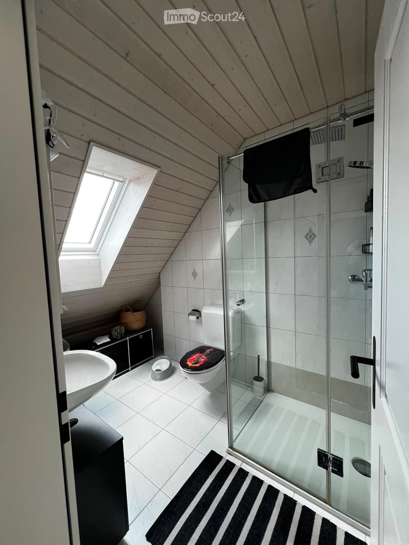 Modern bathroom with white tiles, a glass-enclosed shower, a sink, a toilet with a decorative seat cover, a small black cabinet, and a wicker basket on the cabinet.