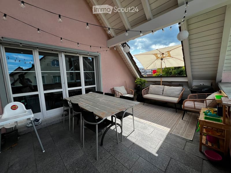 A spacious balcony with a dining table, chairs, a sofa, an umbrella, and a baby chair, offering a view of the city.