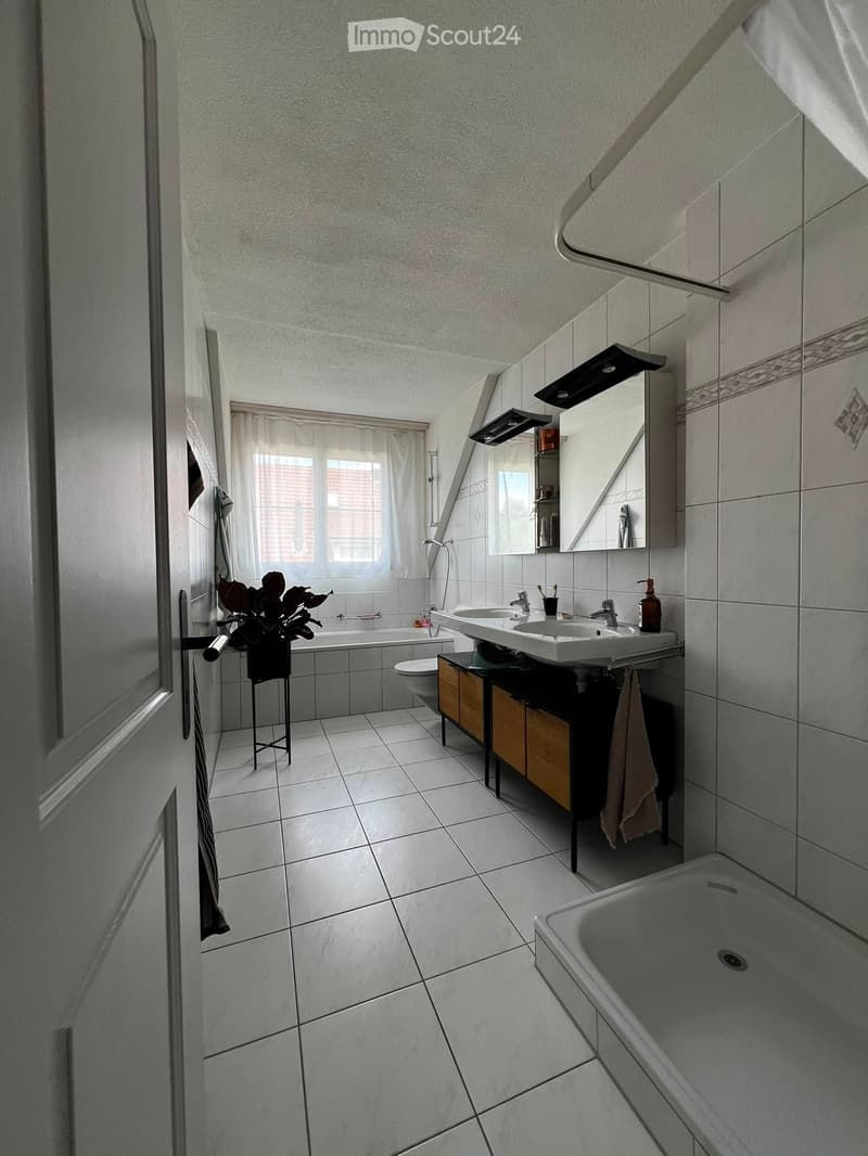 Modern bathroom with white tiles, a bathtub, a double sink, a toilet, a mirror, a small cabinet, and a towel hanging on the side.
