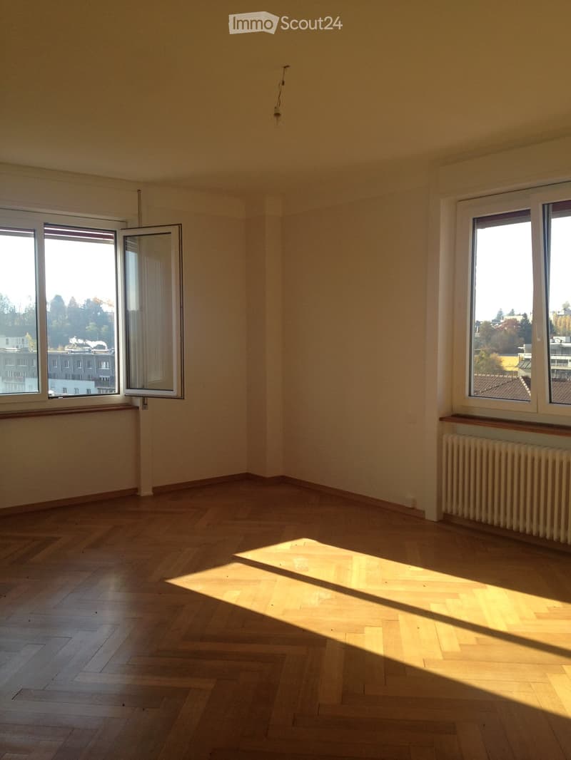 Empty room, parquet floor, white walls, two windows, radiator, and outside view