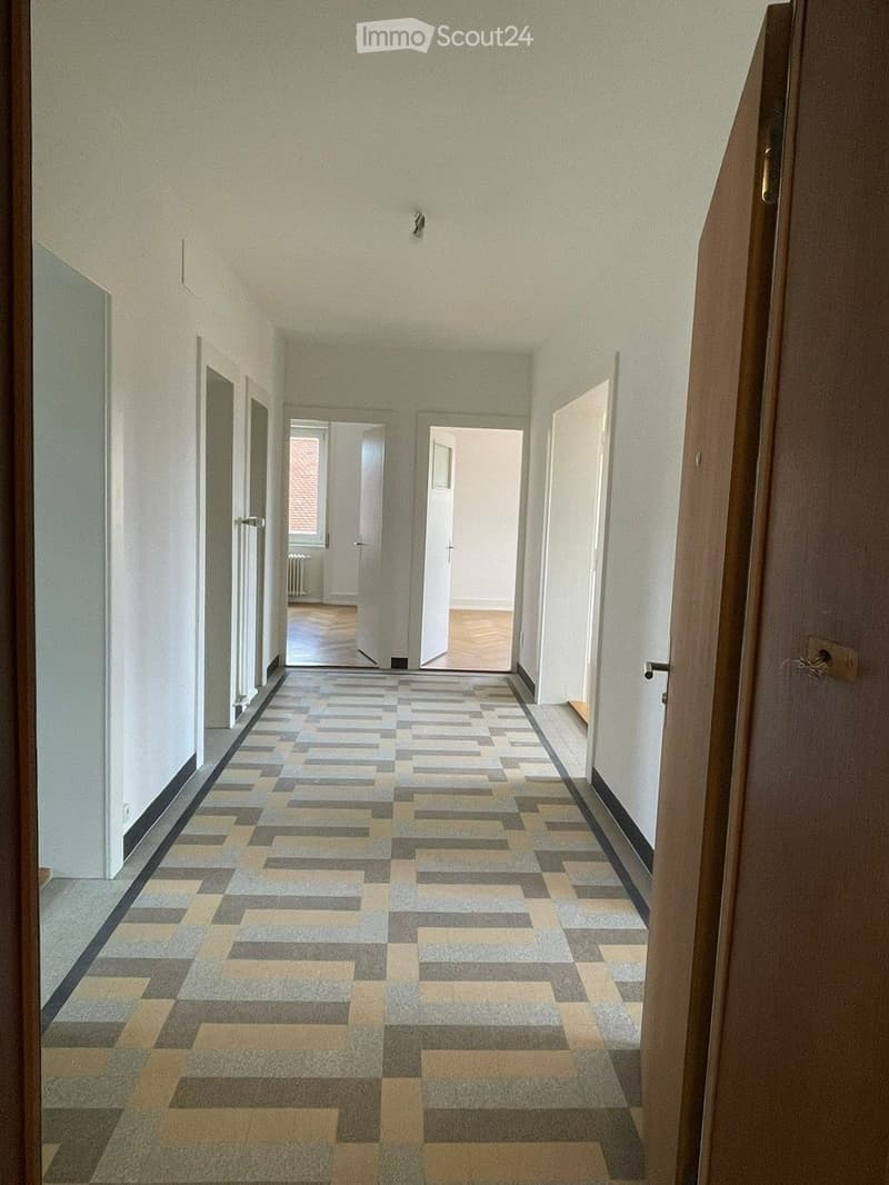 A long, narrow hallway with a geometric patterned carpet in shades of gray and beige, white walls, and several doors leading to other rooms.