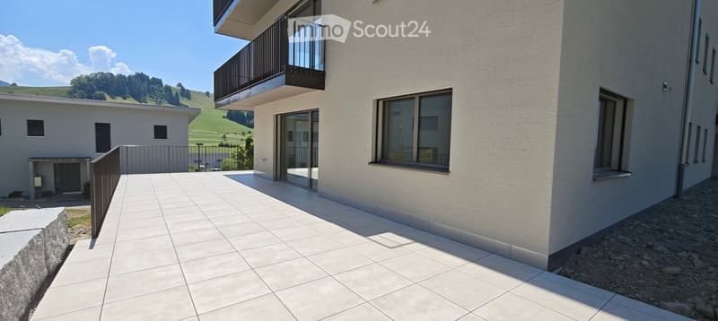 Modern outdoor patio area with white tiled flooring, adjacent to a contemporary building with large windows and a balcony. The background features a scenic green landscape with hills and trees.