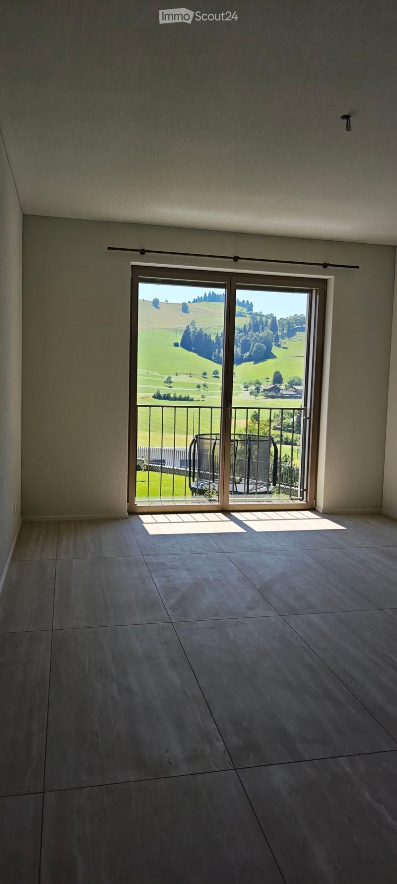 Empty room with large sliding glass doors opening to a balcony, overlooking a green landscape with hills and trees.