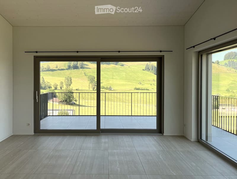Empty room with large sliding glass doors leading to a balcony, offering a scenic view of green fields and trees. The floor is tiled, and the walls are painted in a neutral tone.