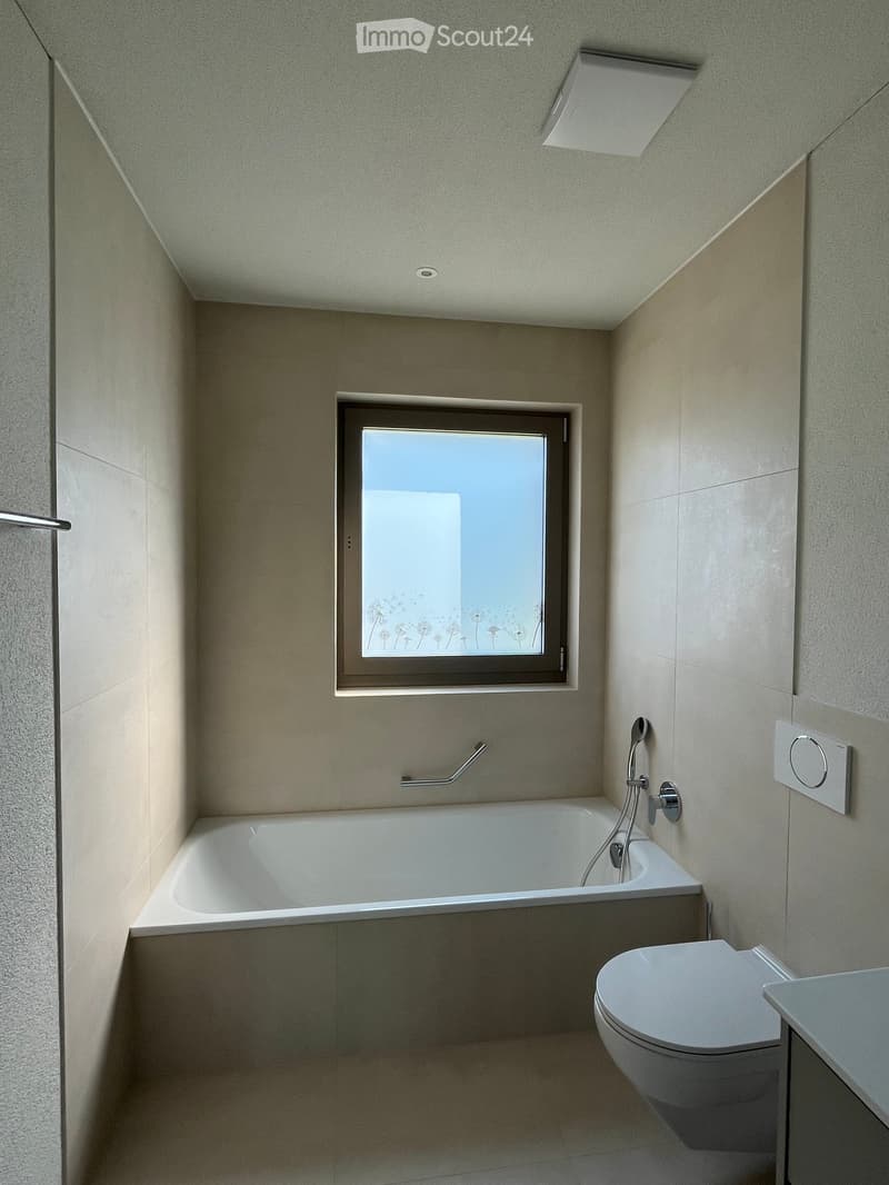 Modern bathroom with a bathtub, toilet, showerhead, and a window with a decorative glass design.