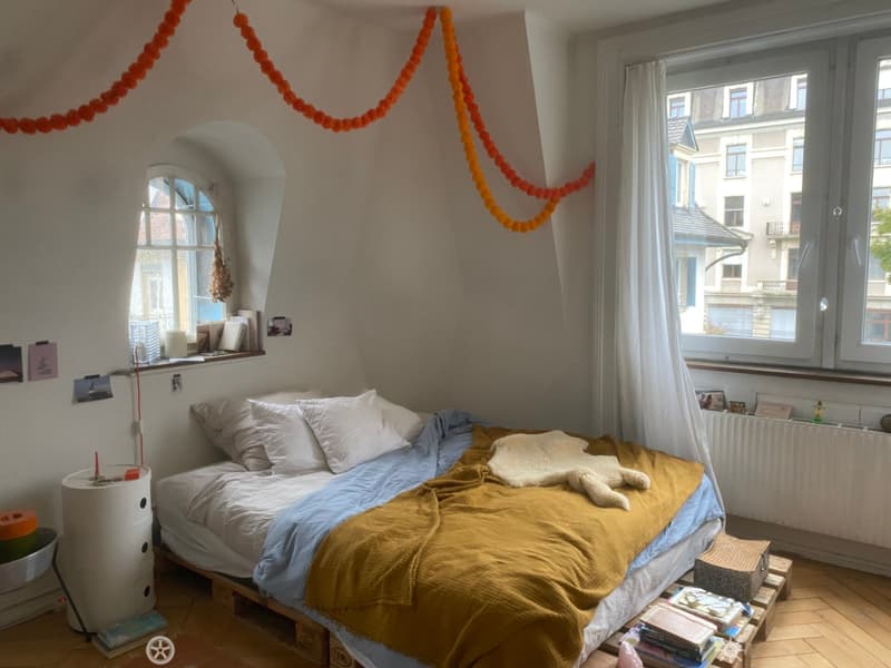 Cozy bedroom with a wooden bed, pillows, blankets, and a small table with books. Large windows with white curtains and a radiator are visible.