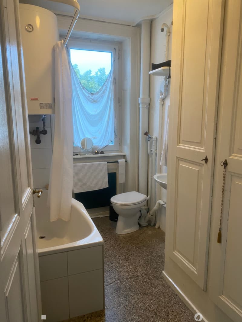Bathroom with a bathtub, toilet, sink, and mirror. The room features a window with a curtain and a towel hanging on the side of the bathtub.