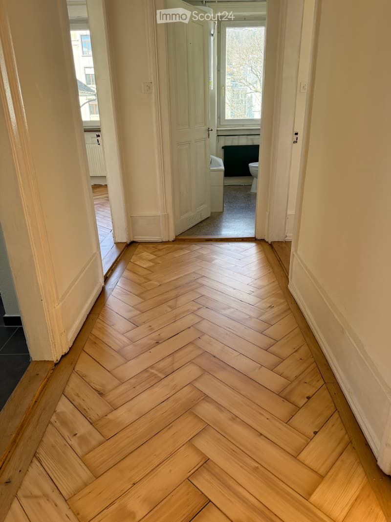 Hallway with herringbone-patterned wooden flooring, white walls, and doors leading to other rooms.