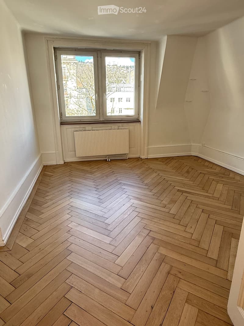 Empty room with herringbone-patterned wooden flooring, white walls, and a large window with a radiator below it.