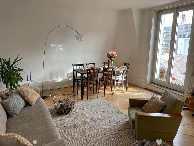 A modern, well-lit living and dining area with a grey sofa, armchair, coffee table, dining table with chairs, floor lamp, and decorative items including a plant, vase, and candles.