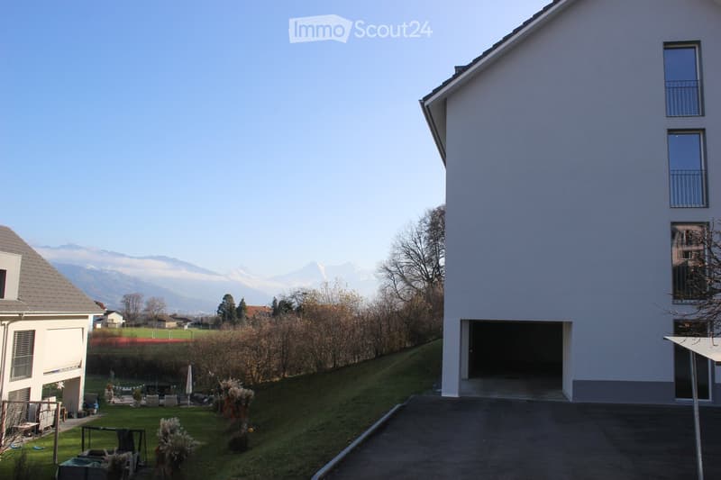 Modern two-story house with white exterior, large windows, and a garage entrance. The surrounding area includes a lawn, trees, and distant mountain views.