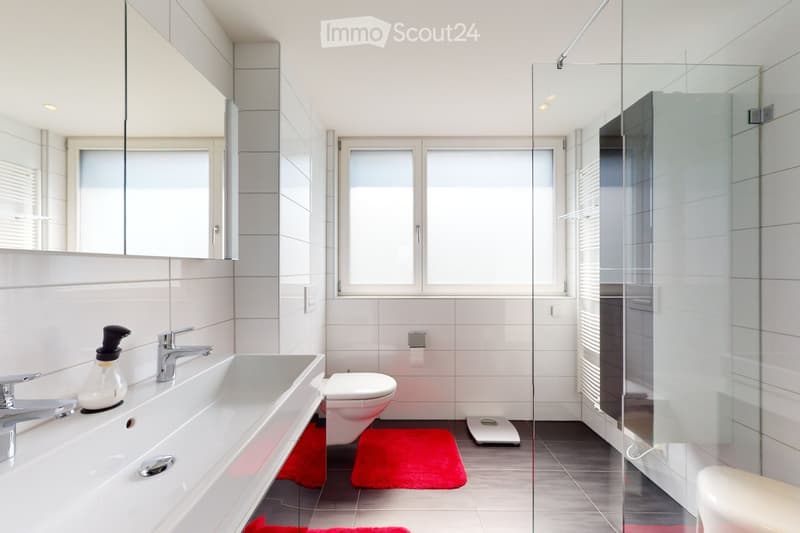 Modern bathroom with white tiles, a large mirror, a sink with a modern faucet, a toilet, a glass-enclosed shower, and a digital scale on the floor.