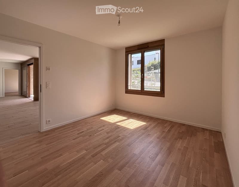 Empty room with wooden flooring, white walls, and a large window with brown frame. Natural light streams in, illuminating the space.