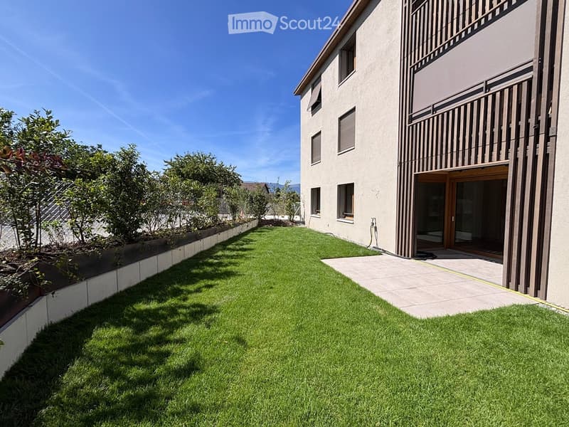 Well-maintained lawn with a raised garden bed, modern house with balcony and large windows, clear blue sky.