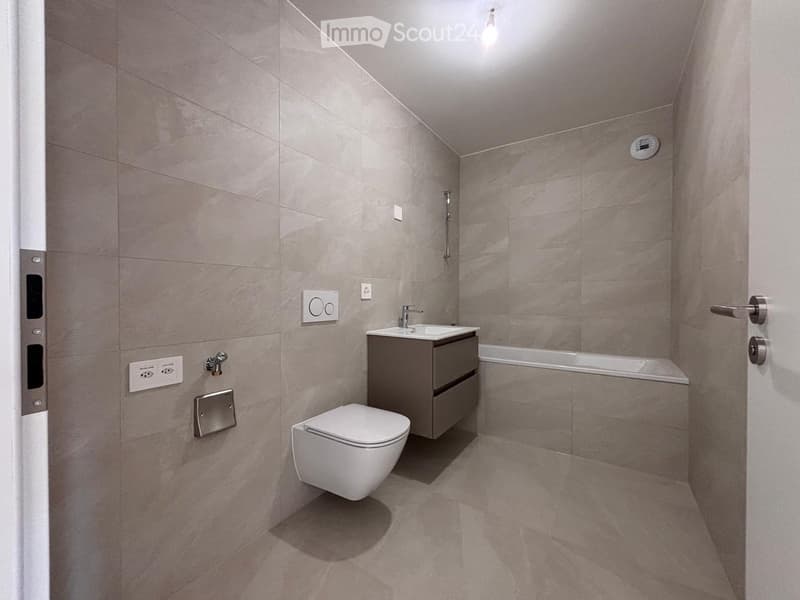Modern bathroom with grey tiled walls and floor, featuring a toilet, sink with a cabinet, and a bathtub.