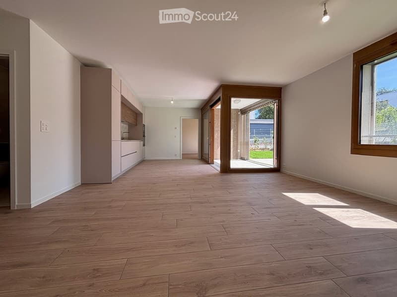 Empty room with wooden flooring, white walls, and large windows. Features a modern kitchen area with cabinets and a doorway leading to an outdoor space.