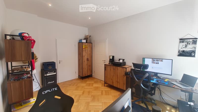 A well-lit room with wooden flooring, a desk setup including a laptop, screen, and chair, a cabinet, a printer, a coffee maker, a guitar amplifier, and a helmet on top of a cabinet.
