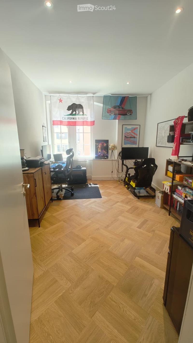 A well-lit room with wooden flooring, a desk setup with a chair, a gaming chair, and shelves with various items. The room features large windows with curtains and artwork on the walls.