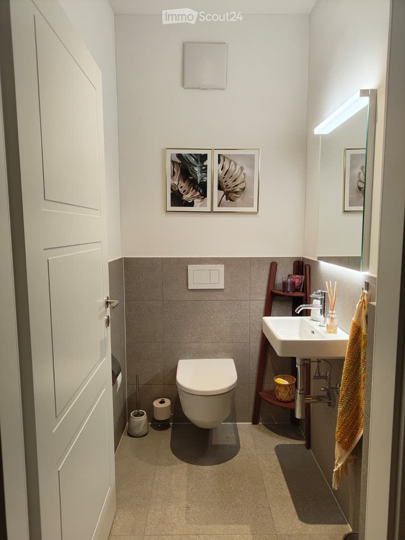 Modern bathroom with a toilet, sink, mirror, and decorative picture frames. Features a towel on a rack and a small wooden shelf with items.