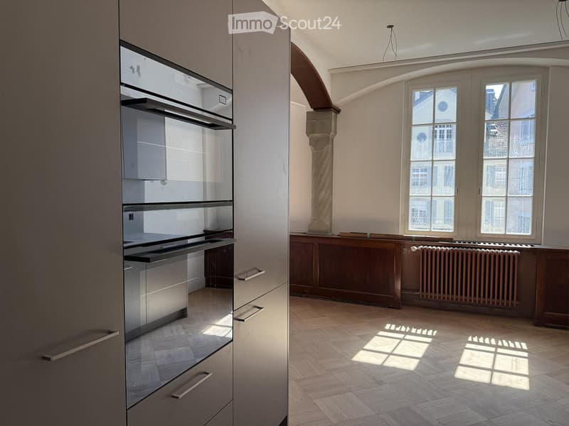 Bright, unfurnished room with wooden flooring, large windows, and a modern kitchen area featuring stainless steel appliances and cabinets.