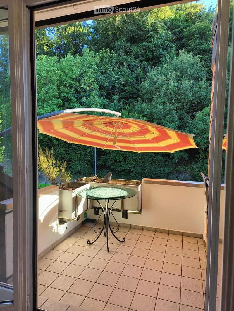 Balcony with tiled floor, glass table, umbrella, and lush green view.