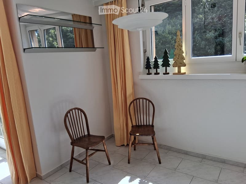 A small, well-lit room with two wooden chairs, a large mirror, a shelf with decorative items, and a table lamp.