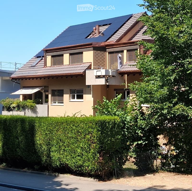 Two-story house with solar panels on the roof, beige and brown exterior, multiple windows, and a well-maintained hedge in the front yard.