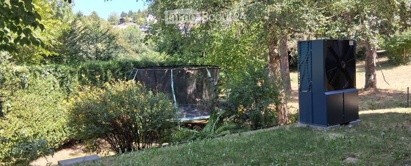 A garden area with a trampoline, surrounded by trees and bushes, and a large black fan unit on the right side.