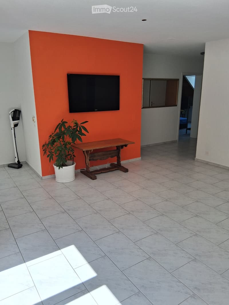Living room with tiled floor, orange accent wall, mounted TV, small wooden table, potted plant, and a vacuum cleaner in the corner.