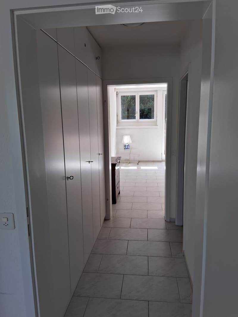 A modern hallway with tiled flooring, white walls, and a built-in cabinet on one side. A small room with a table and lamp is visible at the end of the hallway.