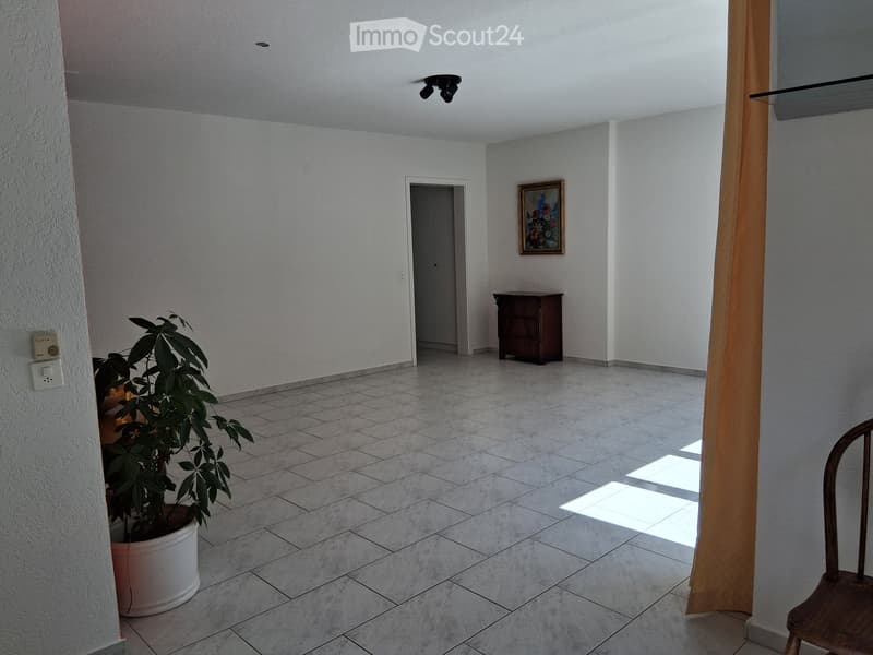 A spacious, well-lit hallway with tiled flooring, a potted plant, a wooden chair, and a small wooden cabinet.