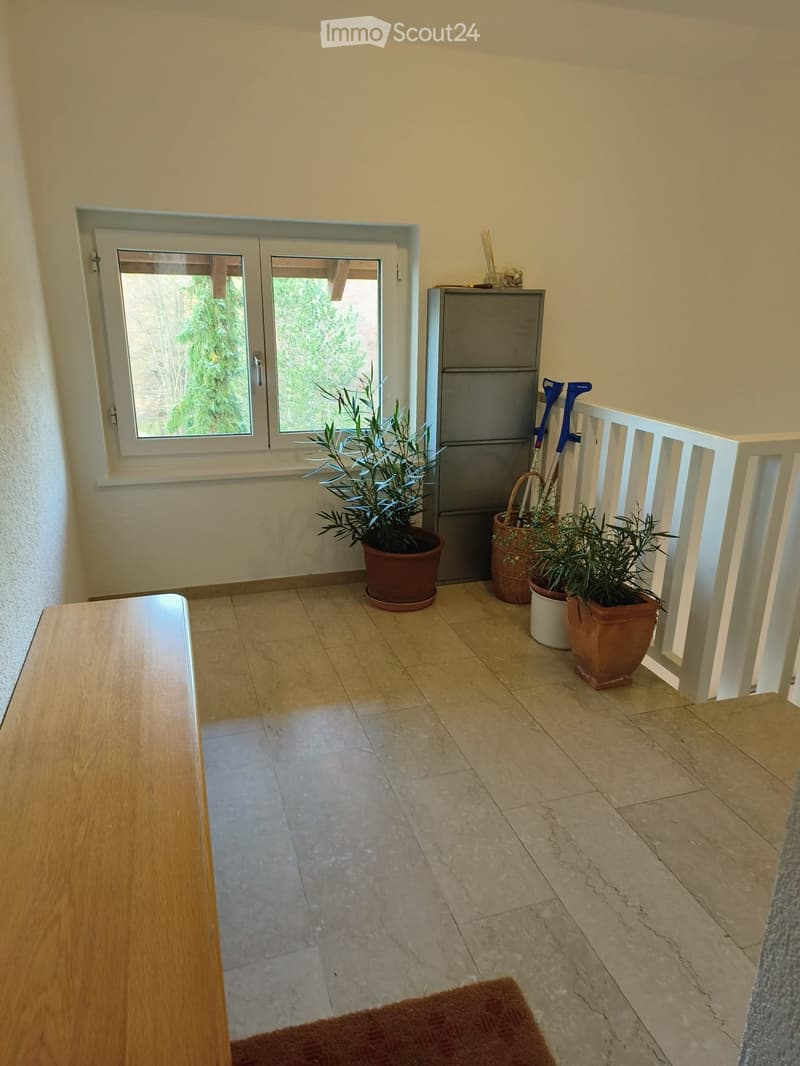 Hallway with tiled floor, white walls, a window with a view of greenery, a small table, potted plants, and a white railing leading to another area.