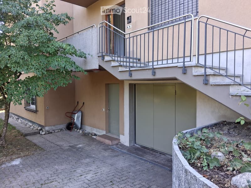 Two-story building with a raised ground floor, accessible via a ramp with metal railings. Features a garage entrance, small garden area, and a tree on the left side.
