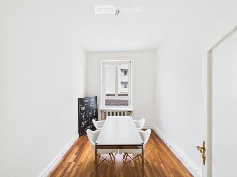 A rectangular white dining table with four white chairs on either side, a blackboard against the wall, white walls and ceiling and wooden flooring, and a window with an outside view.