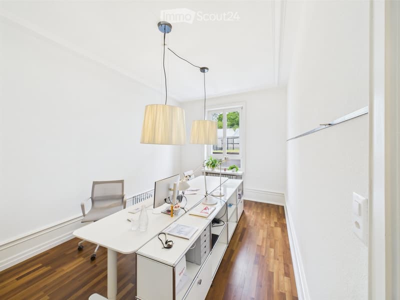 Bright, modern office space with white walls, wooden flooring, a large desk, ergonomic chair, pendant lamps, and a window view of greenery.