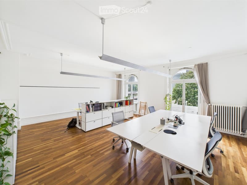 Bright, modern office space with wooden flooring, large white table, several chairs, cabinet with books, laptop, and plants near the window.
