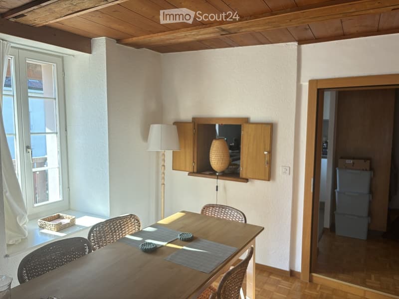 Bright dining area with wooden table, wicker chairs, a floor lamp, and a mirror on a wooden shelf. The room has wooden flooring and white walls, with a window offering natural light.