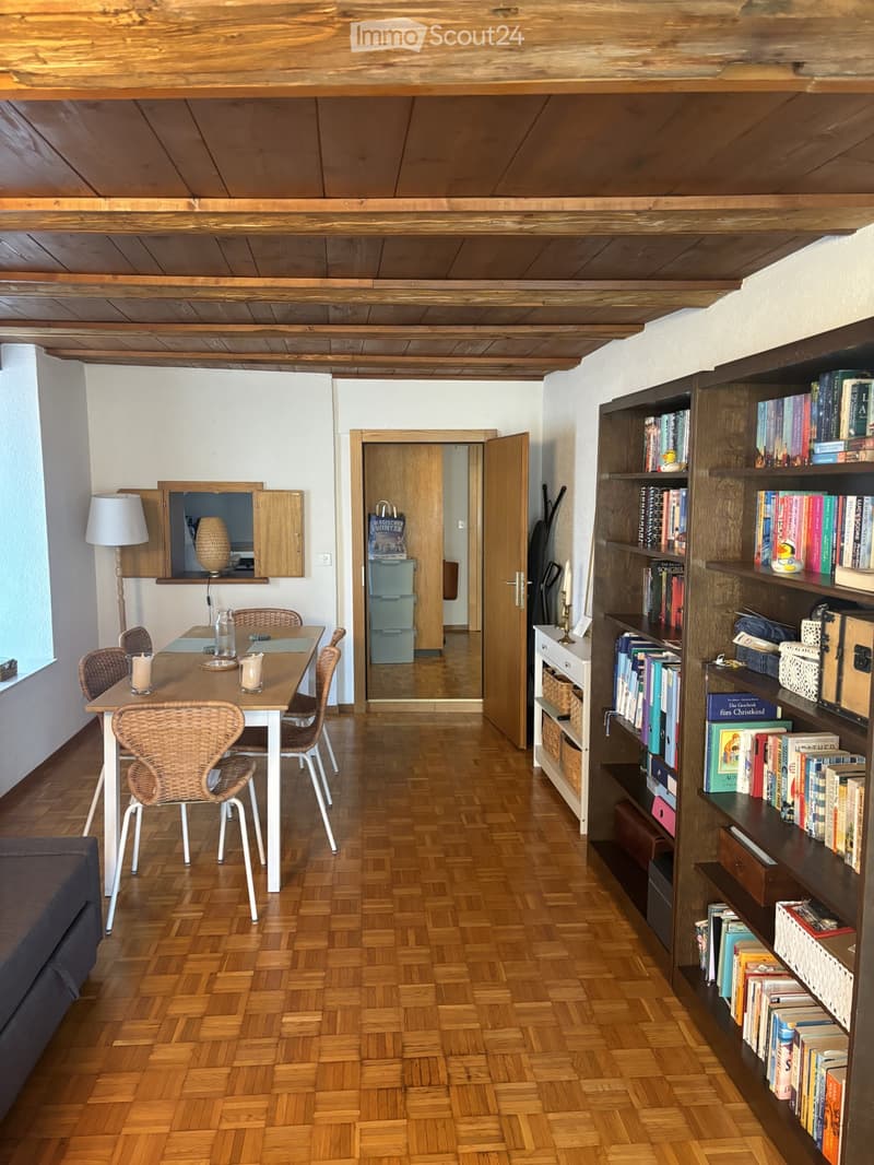 A cozy, well-furnished room with wooden floors, a large bookshelf filled with books, a dining table with chairs, a lamp, and a cabinet. The room features exposed wooden beams on the ceiling and a doorway leading to another space.