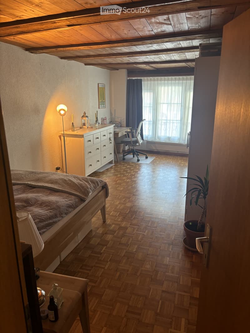 Cozy bedroom with wooden flooring, a bed with a fur throw, a desk with a chair, a drawer unit, a floor lamp, and a potted plant near the door.