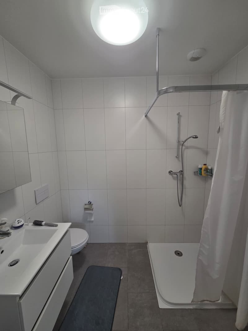 Modern bathroom with white tiles, a bathtub with a shower curtain, a sink with a mirror, a toilet, and a shower shelf with toiletries.