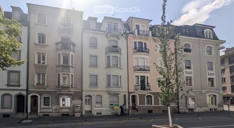 A row of multi-story residential buildings with balconies, large windows, and classic architectural details, situated on a street with a tree and a few pedestrians.
