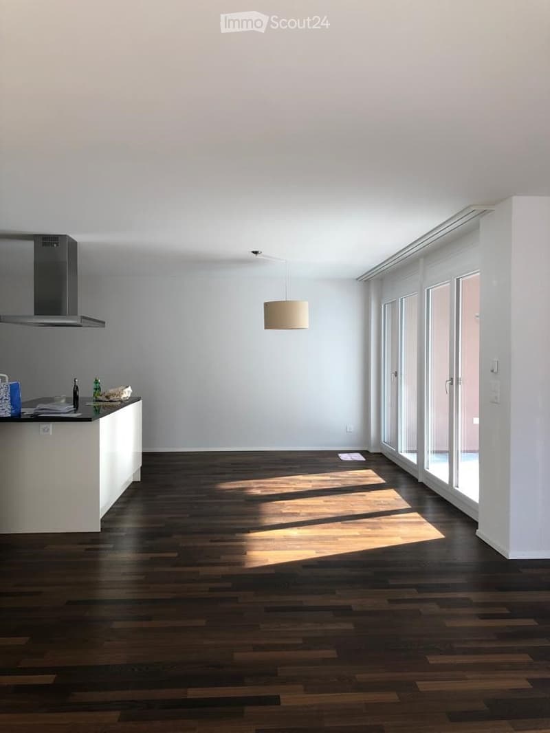 Empty room with dark wooden flooring, white walls, and large windows allowing natural light to enter. Minimal decor with a small counter area visible.