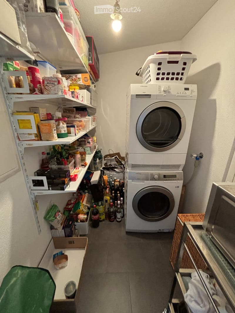 A small, well-lit room with a washing machine and dryer stacked vertically, shelves filled with food items and household supplies, and a laundry basket on top of the dryer.
